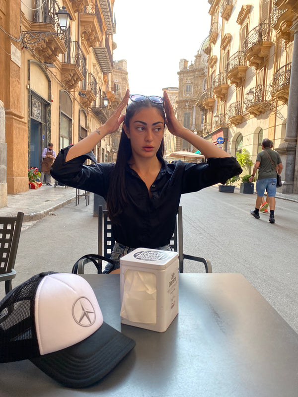 Girl sitting in street café in Palermo, Sicily with Verona Concepts Peace Trucker Cap on the table.